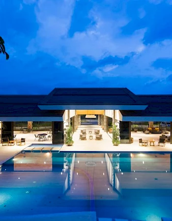 An outdoor swimming pool at night in front of a modern resort building under a vivid blue sky.