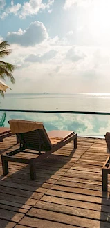 Oceanfront resort deck with lounge chairs and umbrellas beside a reflective pool, with palm trees and calm sea at sunset.