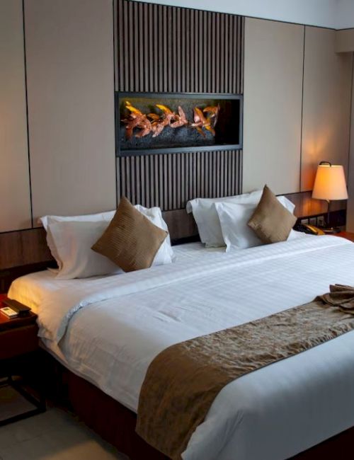 A modern hotel room with a made bed, pillows, bedside lamps, and a wall fireplace featuring burning logs.
