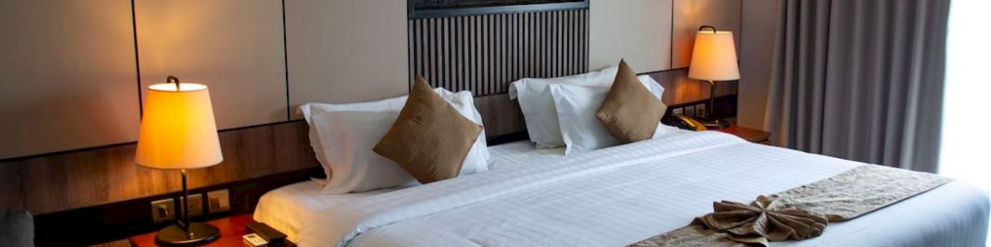 A modern hotel room with a neatly made bed, warm bedside lamps, and a lit fireplace feature on the wall.