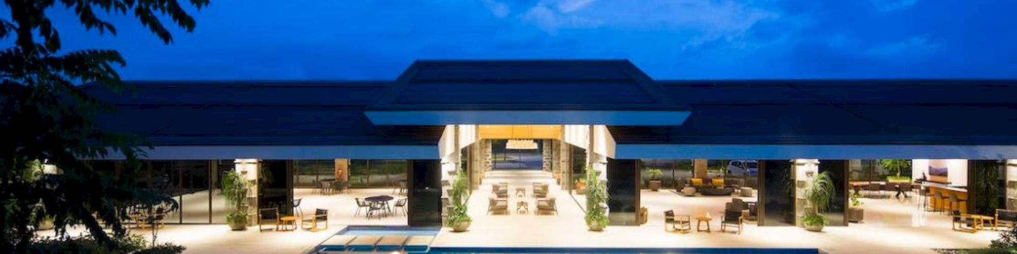 A modern hotel courtyard at dusk with a lit swimming pool, lounge chairs, and palm trees under a blue sky.