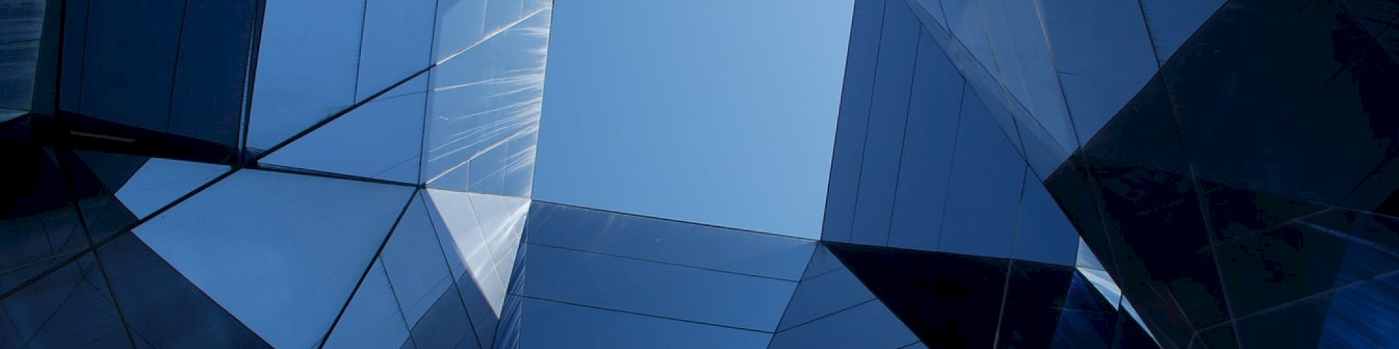 An upward view of a modern blue glass building with angular geometric panels and sharp reflections.