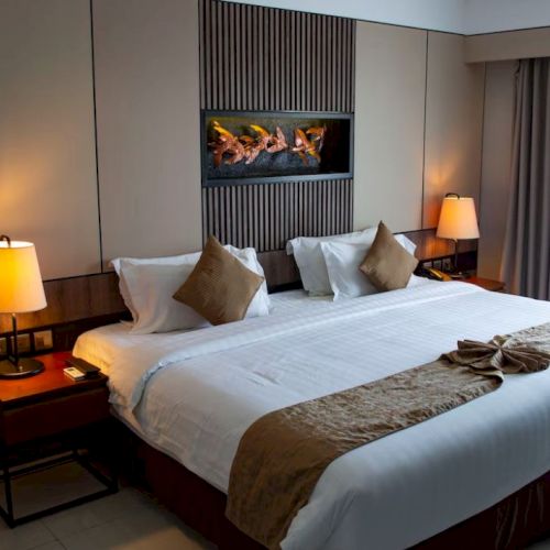 A modern hotel room with a neatly made bed, bedside lamps, and a stone fireplace glowing behind the headboard.