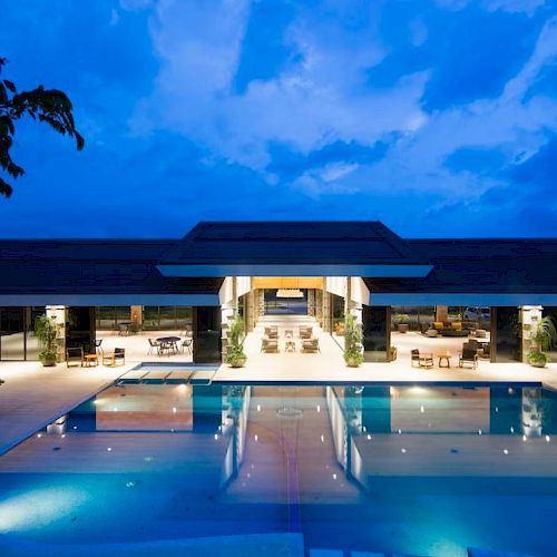 A modern hotel at dusk with a lit swimming pool and lounge seating under a blue sky, framed by palm trees.