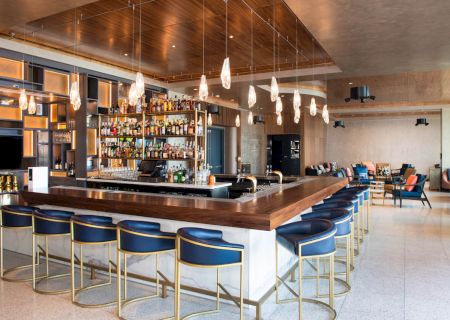 A modern bar lounge with a wooden counter, hanging pendant lights, stocked shelves, and blue high stools beside seating.