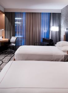 A modern hotel room with two neatly made beds, a wall-mounted TV, desk space, and dark curtains framing the window.