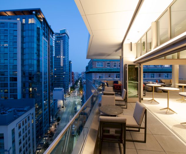 Rooftop terrace with lounge chairs beside sliding windows, overlooking tall city buildings and streets at blue-hour dusk.