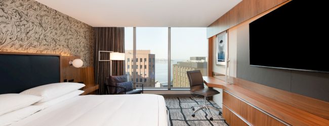 Modern hotel room: bed, desk chair, wall-mounted TVs, patterned wallpaper, and a city view through large windows.