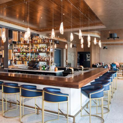 A modern hotel bar with a long wooden counter, bottle-lined shelves, hanging lights, and blue lounge seating.