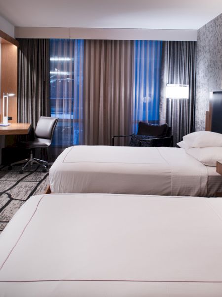 A modern hotel room with two beds, wall-mounted TV, desk area, and large curtains with blue light filtering through the windows.