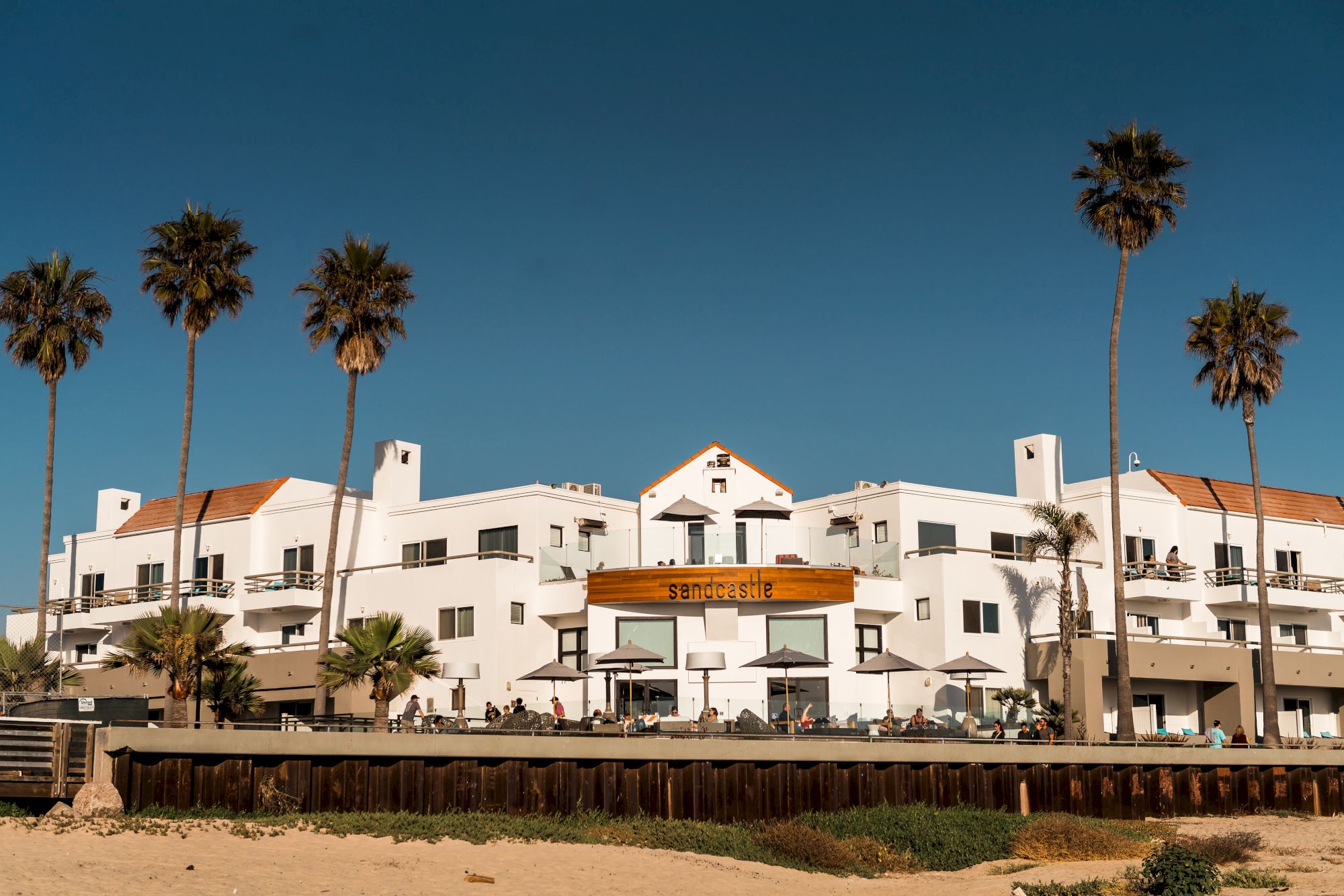 White coastal buildings with orange-tiled rooftops line a sandy beach, palm trees stand tall, a wooden barrier divides the shore, calm blue sky.
