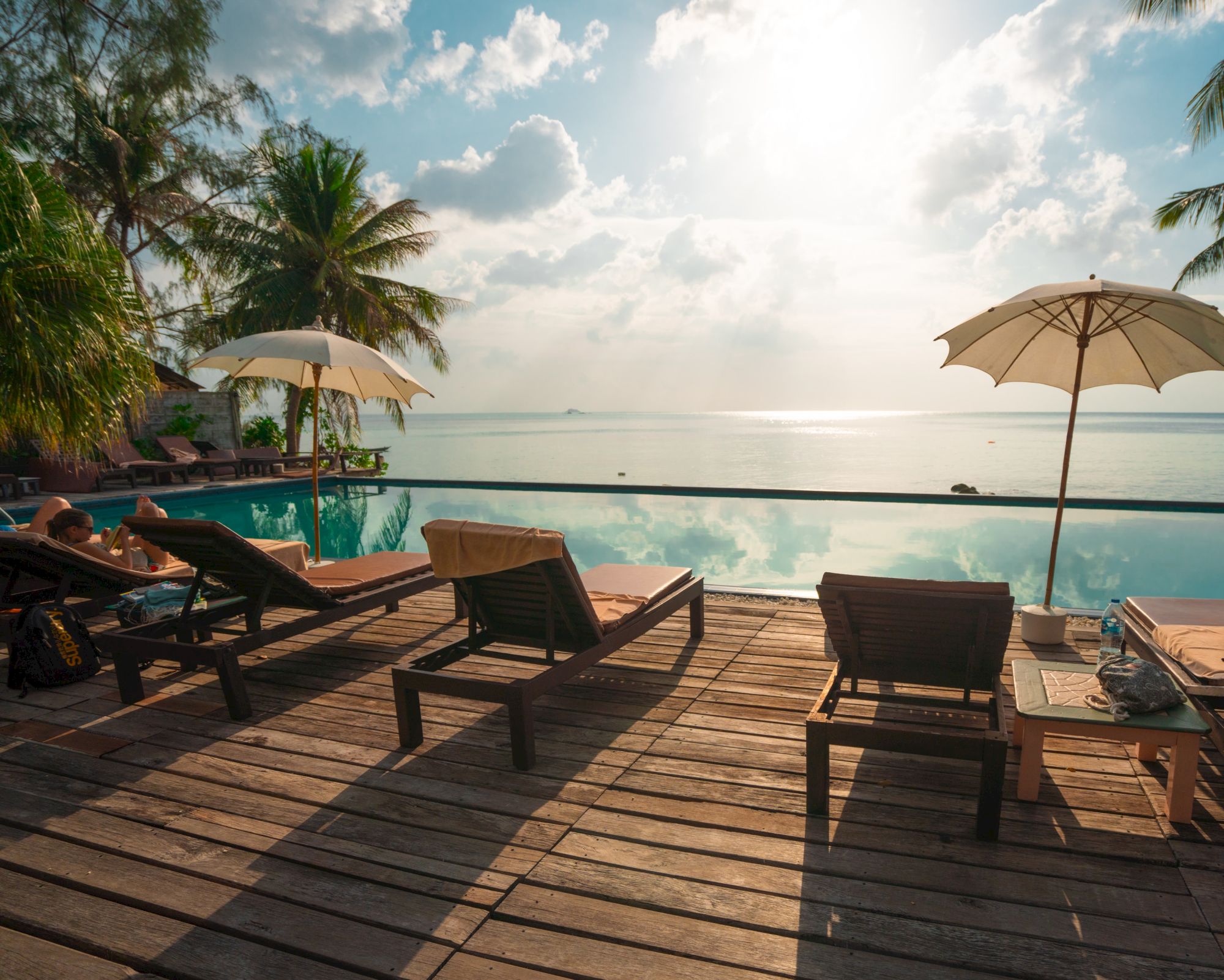 A tropical beachfront resort with a pool, sun loungers and umbrellas, palms, and calm ocean under bright clouds.