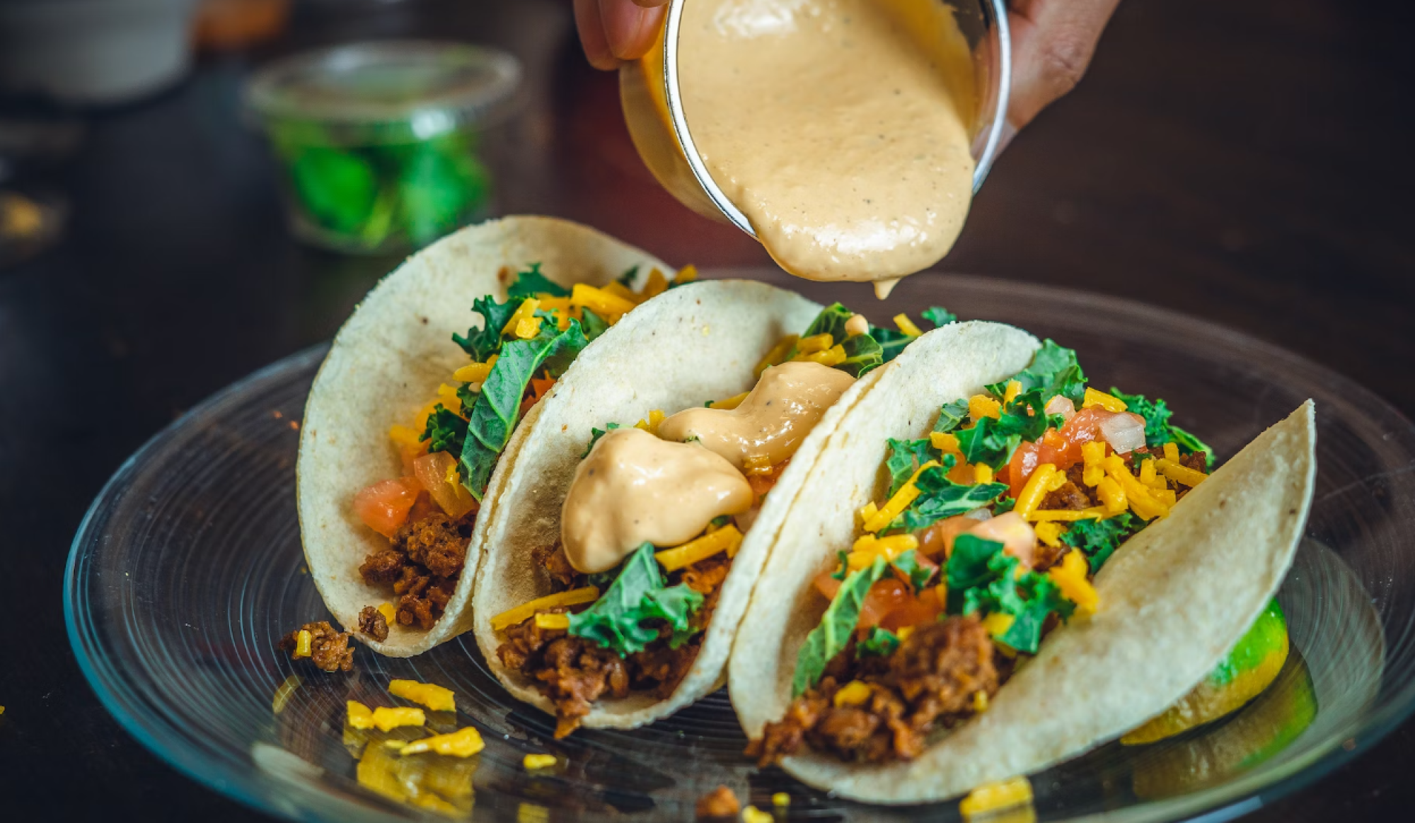Tacos on a plate with shredded veggies and meat, with creamy sauce being poured over them.