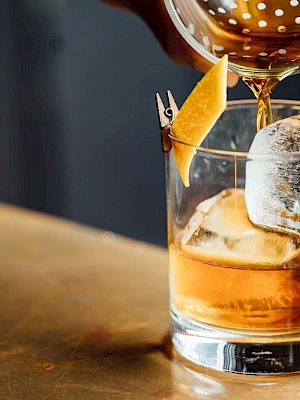 An amber cocktail is poured into an ice-filled glass, topped with a garnish and held by a second tipped drink.