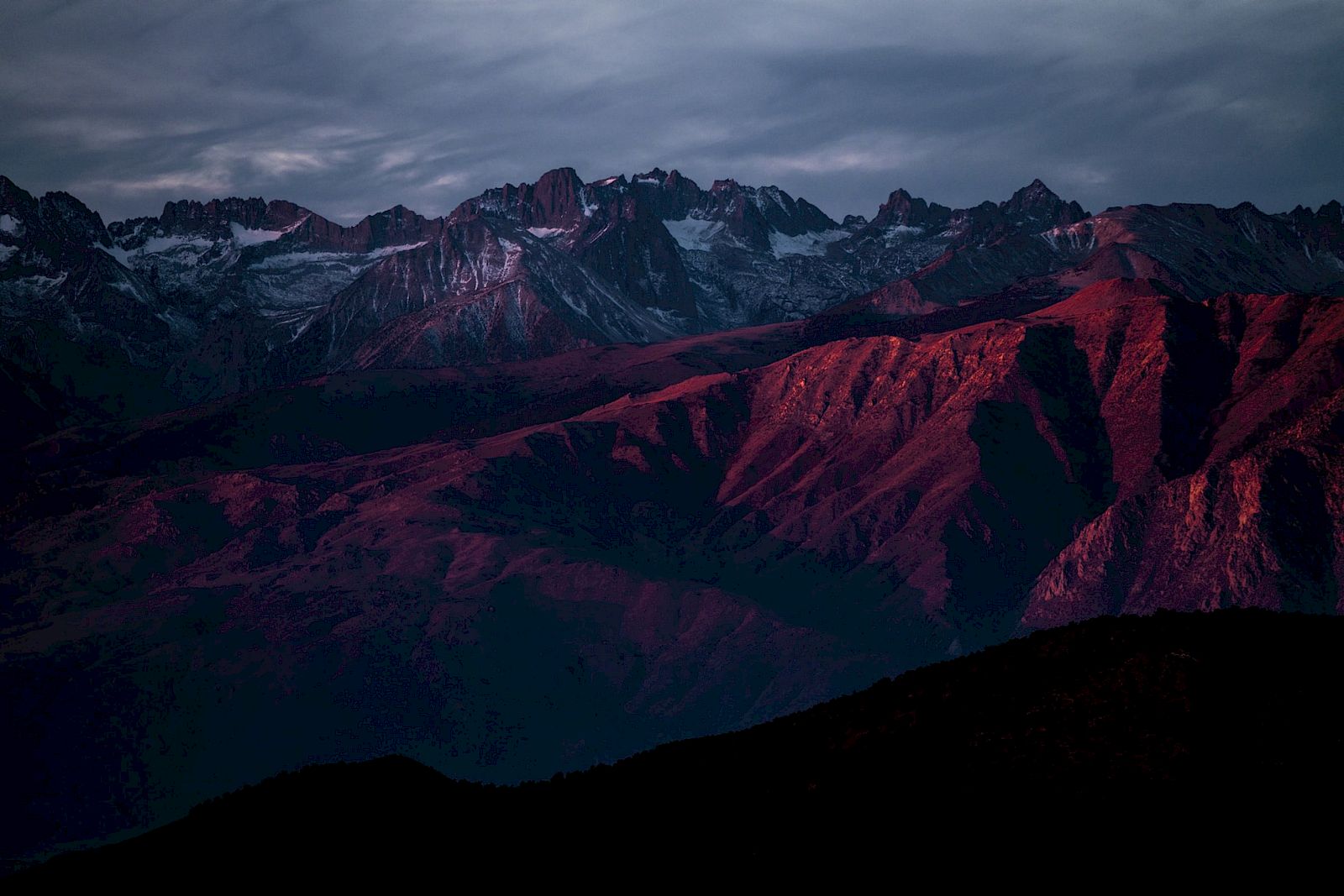 Dramatic mountain range at dusk with snowy peaks, dark valleys, and crimson-lit ridges under cloudy stormy skies.