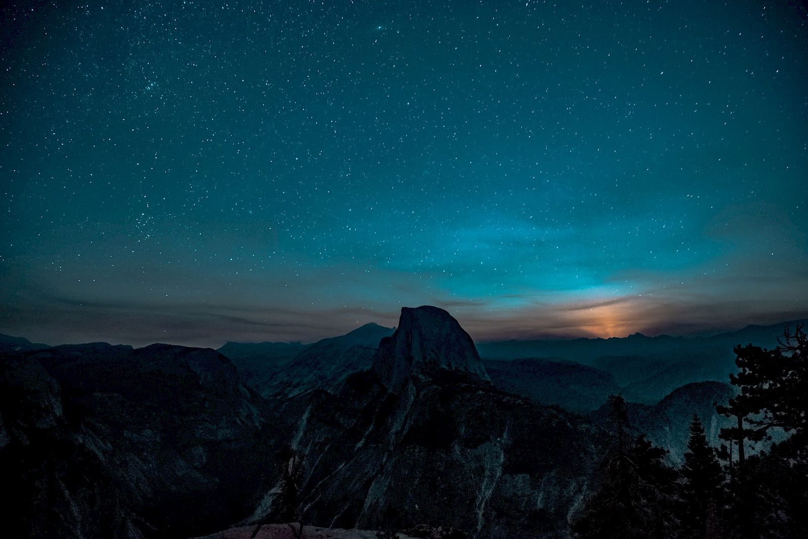 A star-filled night sky over dark mountain peaks, with the Milky Way arcing above and a glowing horizon.