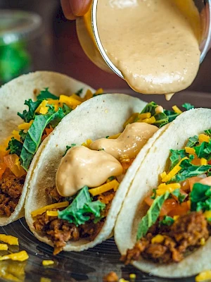 Tacos on a plate being topped with creamy sauce, chopped veggies, and shredded cheese.