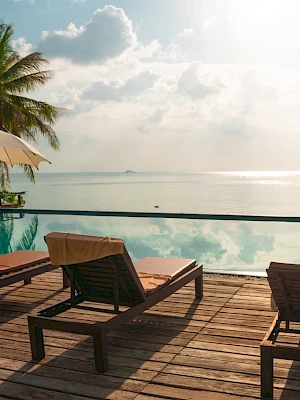 Tropical beachfront resort deck with sun loungers, umbrellas, a small pool, and palm trees overlooking the ocean.