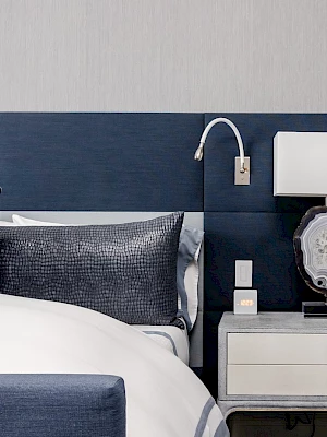A modern bedroom with a made bed, navy headboard, and a nightstand holding a lamp, phone, and decorative display plate.