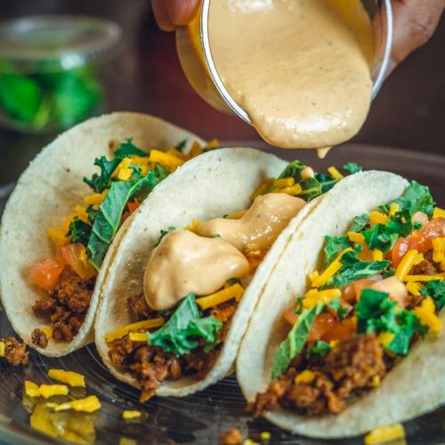 Three tacos on a plate with chopped toppings, with cheese sauce being drizzled over them.