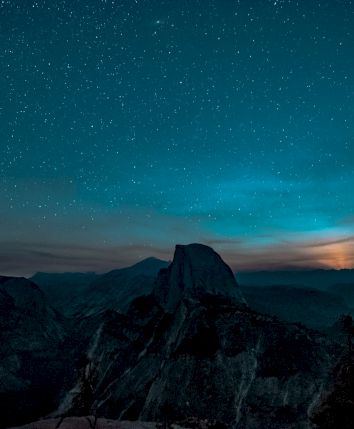 A clear night sky filled with stars over dark mountain peaks, with a faint bluish aurora-like glow near the horizon.