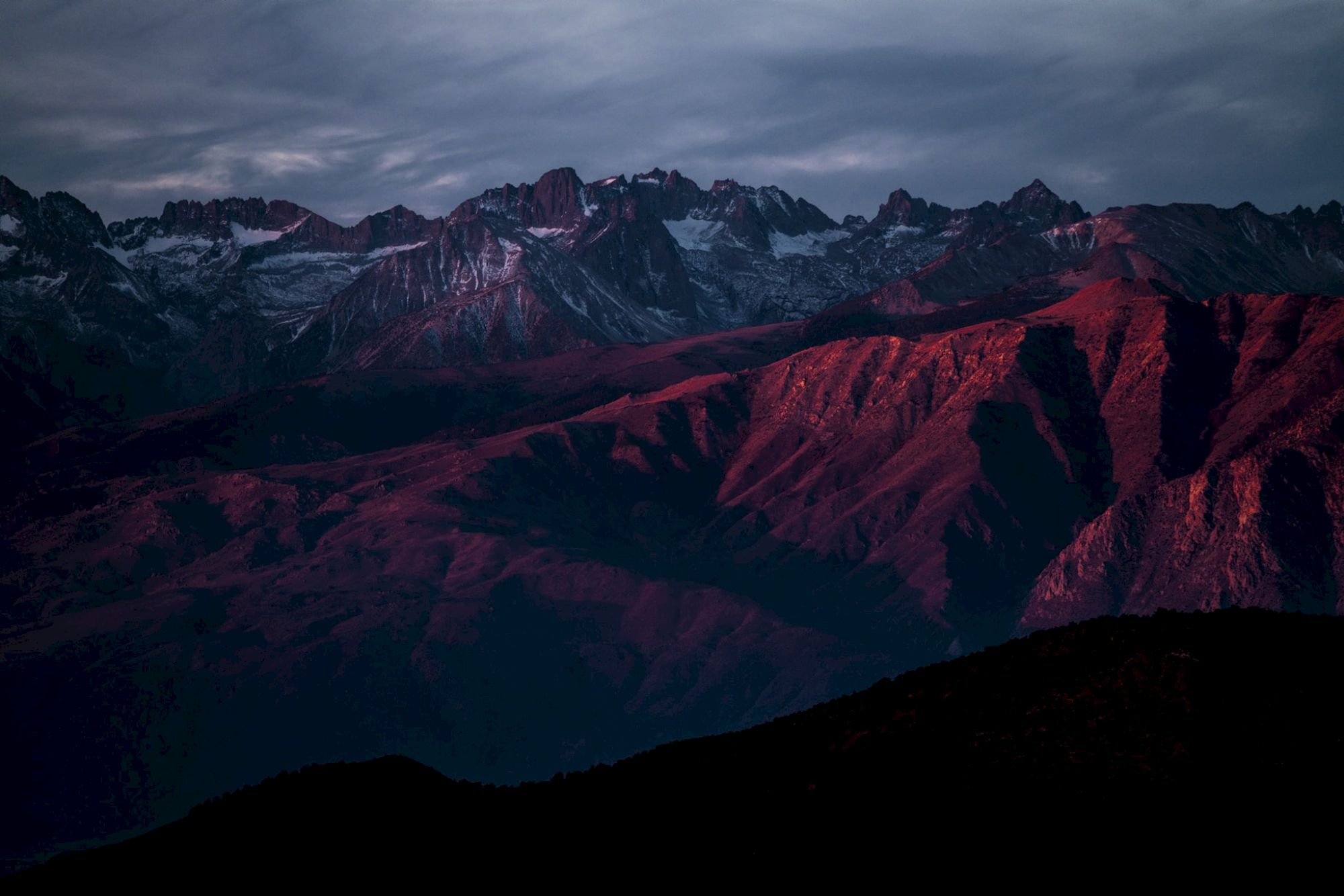 Moody mountain range at dusk, snow-dusted peaks glowing red with dramatic clouds overhead.