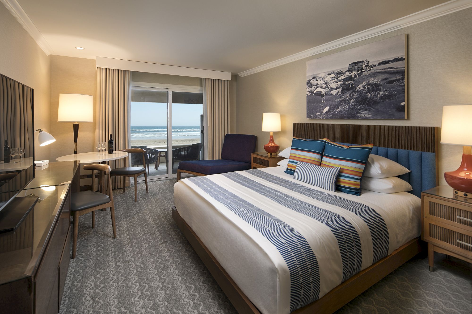 A stylish hotel room with a king bed, blue striped bedding, two nightstands, a desk, a TV, and a balcony overlooking the ocean. End.