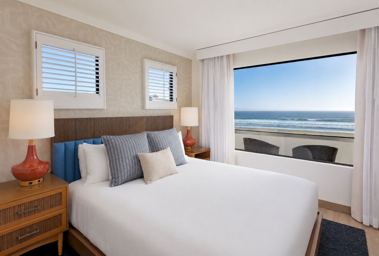 Cozy hotel bedroom with ocean view, made bed, blue pillows, bedside lamps, and curtains framing the sea.