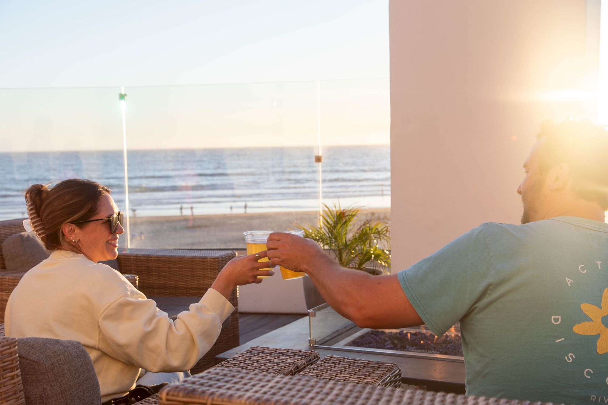 A couple clinks orange drinks on a seaside balcony at sunset, relaxing together with the ocean waves behind them.