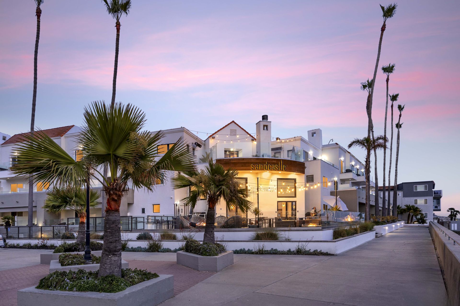 A resort courtyard at dusk with tall palm trees, string lights, and a &ldquo;sandcastle&rdquo; building entrance.