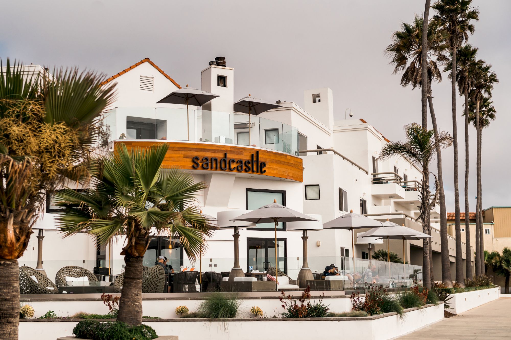 A white seaside hotel with orange sign, outdoor seating, umbrellas, palm trees, and a bright, sunny sky.