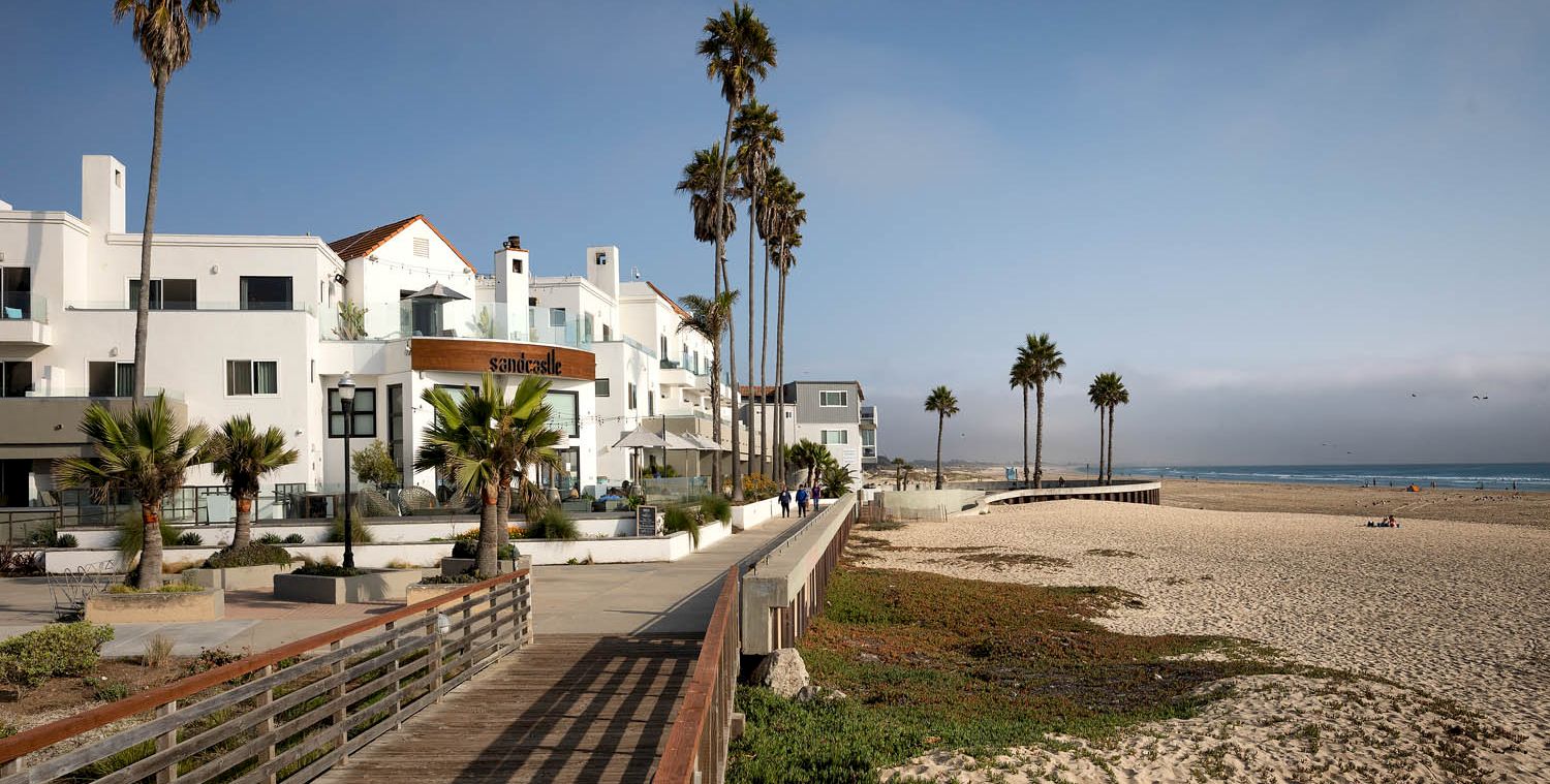 Residential beachfront scene with white modern homes, palm trees, a boardwalk, and a sandy shore meeting the calm ocean, under a clear sky.