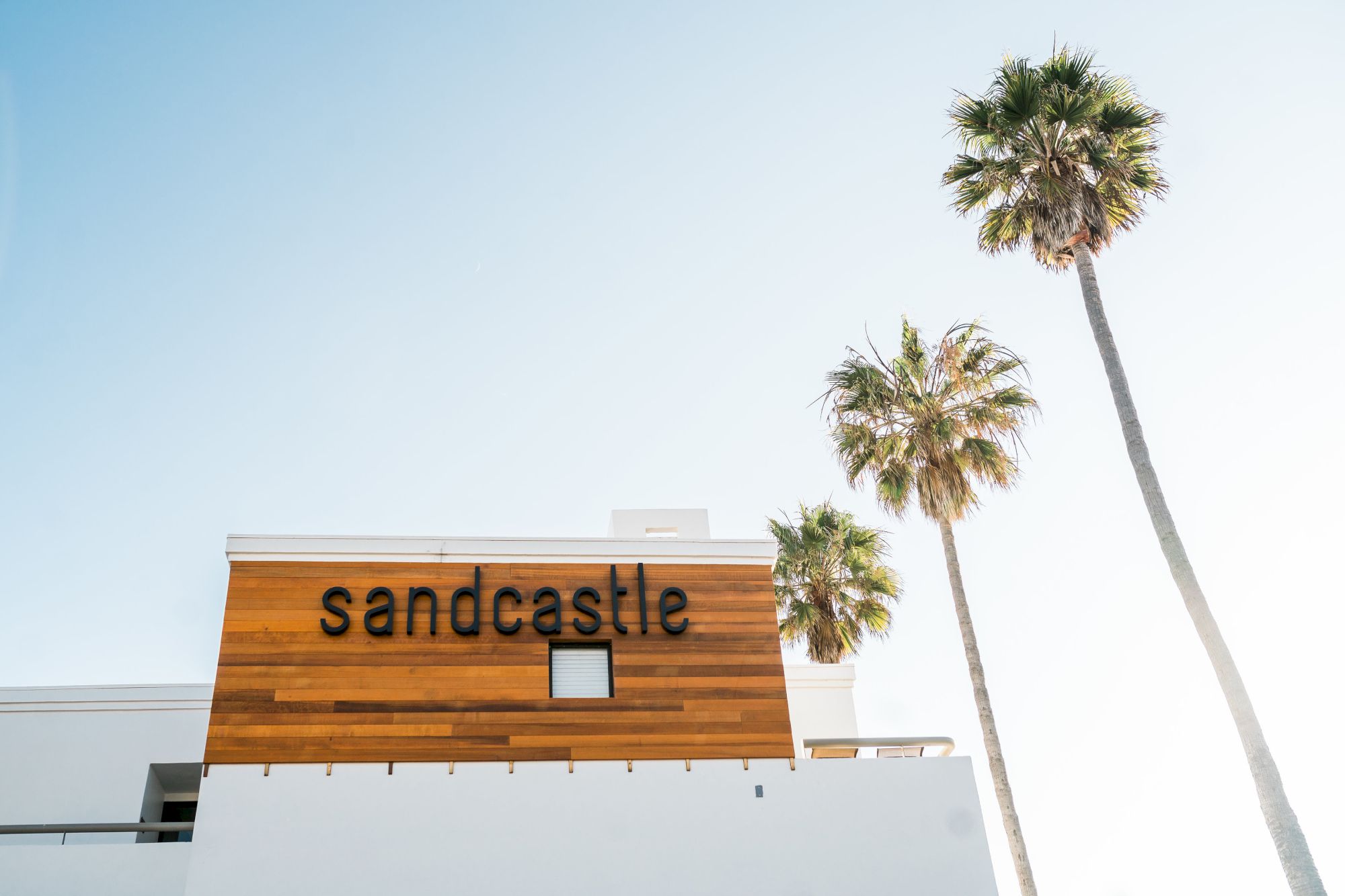 A stylish sign reading &ldquo;sandcastle&rdquo; on a white building, with tall palm trees against a bright blue sky.