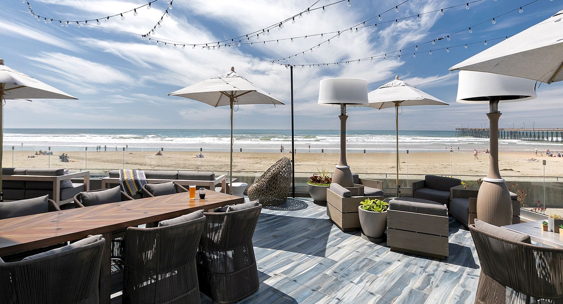 A beachside outdoor dining area with umbrellas, string lights, and a view of the ocean and sandy beach.