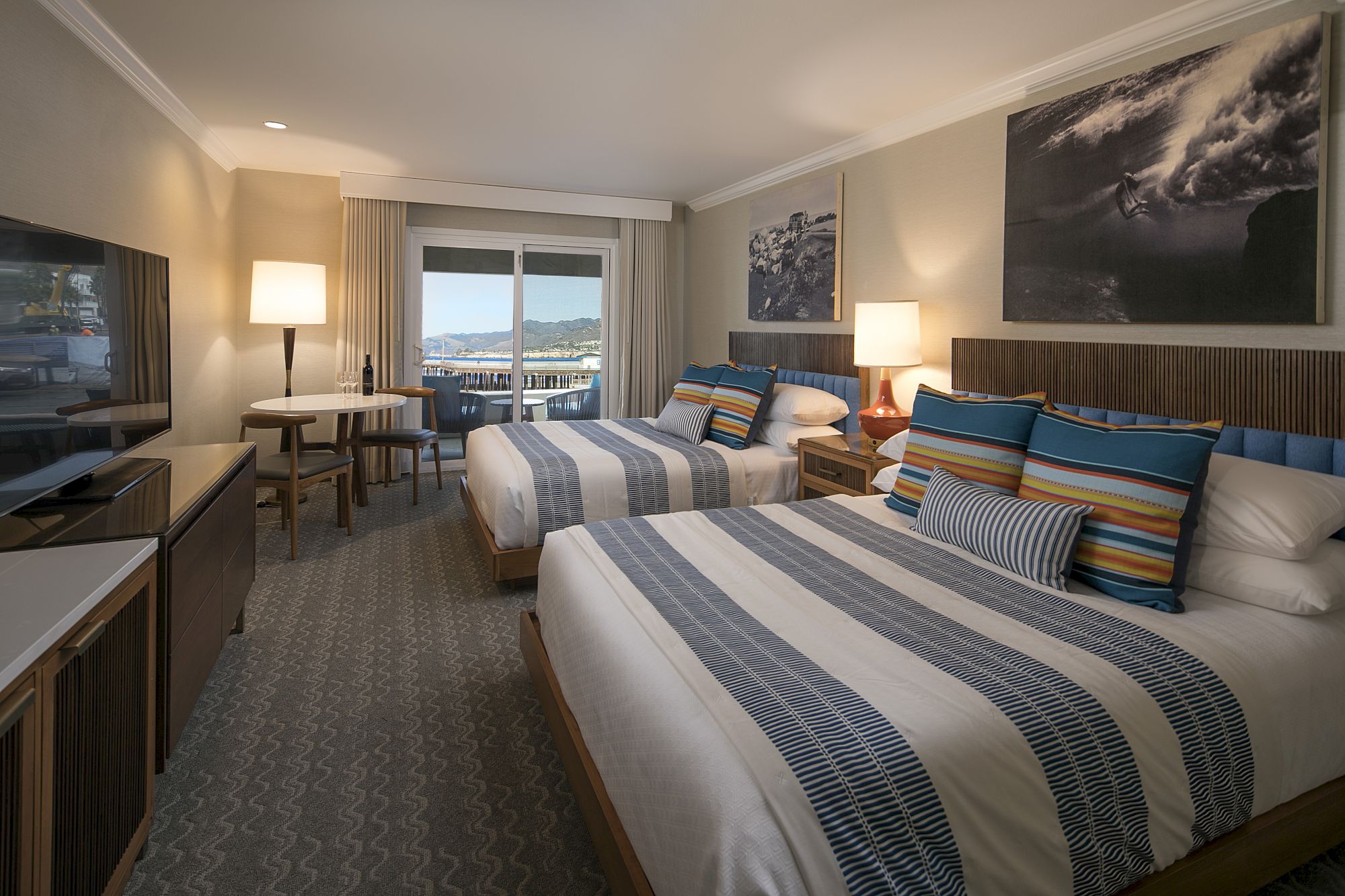 A bright hotel room with two neatly made beds, colorful pillows, bedside lamps, wall art, and a view through large windows.