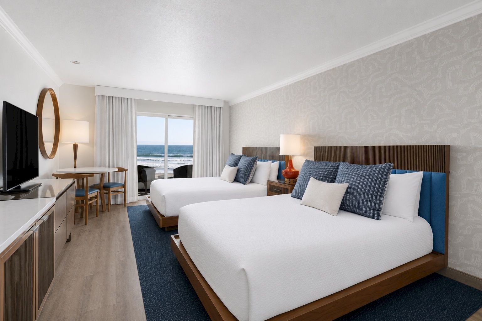 A modern hotel suite with two beds, blue pillows, lamps, and ocean waves visible through a window.