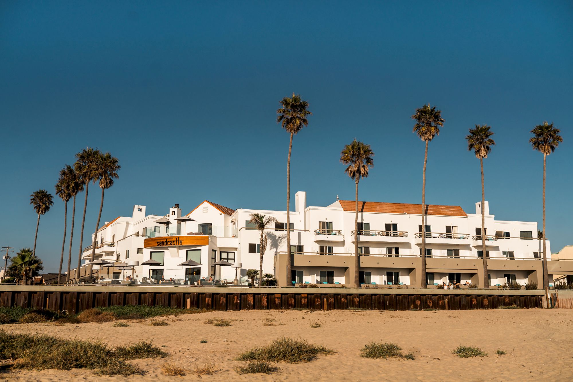 A beachfront hotel with white buildings and orange rooftops, surrounded by tall palm trees under a clear blue sky.
