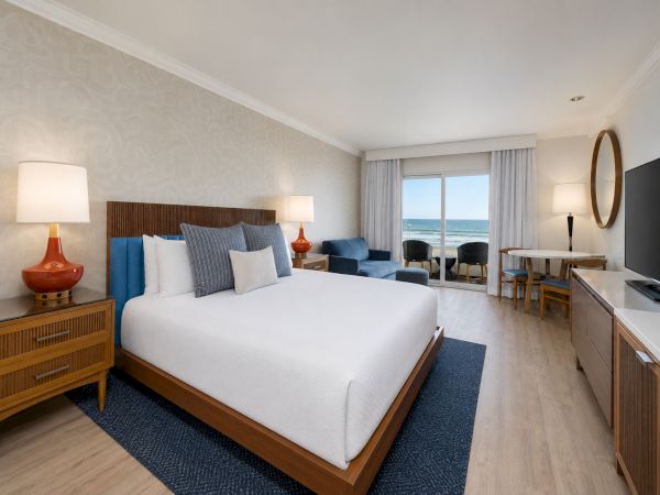 Bright seaside hotel bedroom with a large bed, blue seating, lamps, and a window showing the ocean.