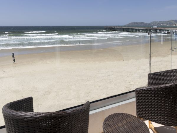 A sunny beach with rolling waves and a long pier, viewed from outdoor wicker chairs in the foreground.