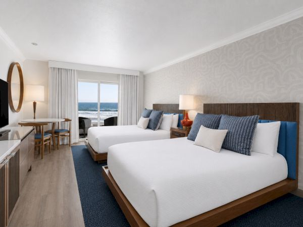 A bright hotel room with two beds, blue pillows, a TV and ocean view through floor-to-ceiling windows.