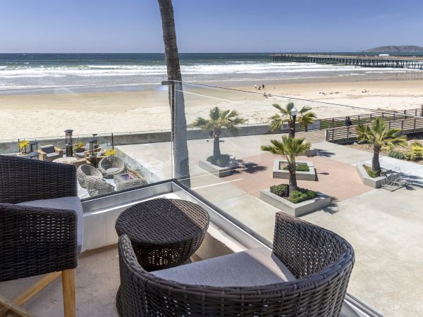 A beachfront view from a balcony with wicker chairs, a small table, palm trees, sandy shore, and a pier stretching into the ocean.