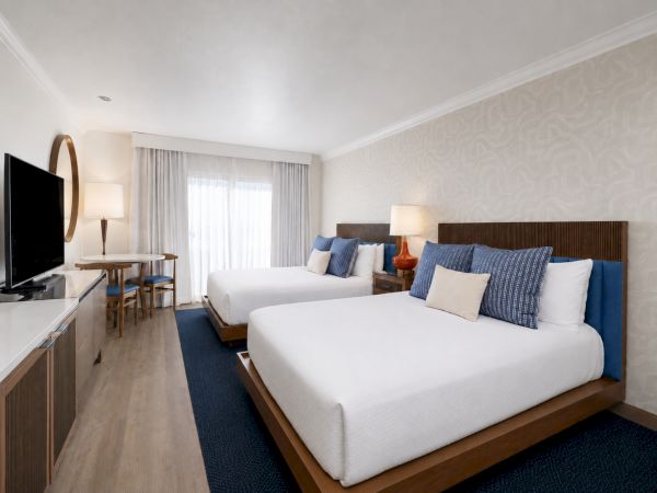 A modern hotel room with two neatly made beds, blue accents, bedside lamps, and a flat-screen TV on a dresser.