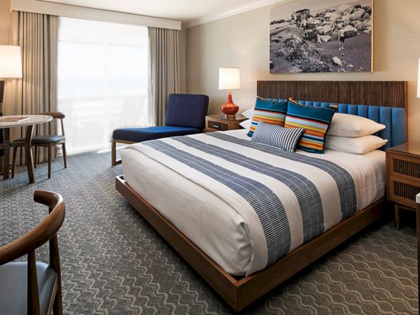 A stylish hotel room with a king bed, blue accents, desk, seating area, lamps, and a large window with curtains.