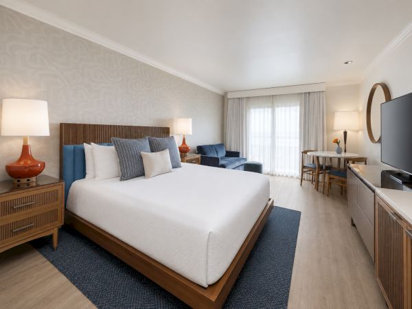 A modern hotel-style bedroom with a large bed, bedside lamps, seating area, floor-to-ceiling curtains, and a wall-mounted TV.