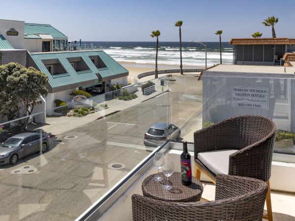Oceanfront resort view with palm trees, a parking lot, and a balcony table, plus a &ldquo;Pismo On the Beach&rdquo; vacation rentals sign.