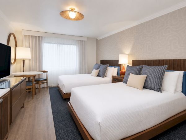 A modern hotel bedroom with two white beds, blue pillows, bedside lamps, a window, and a flat-screen TV on the left.