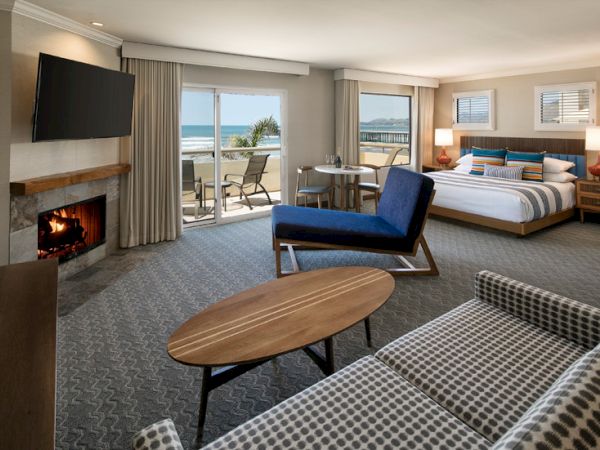 Cozy hotel suite with a fireplace, sofa, chairs, coffee table, and a balcony overlooking the ocean; lights on, smooth textures, relaxing vibe.