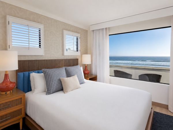 A cozy hotel bedroom with an ocean view, large bed, lamps, blinds, and sheer curtains.