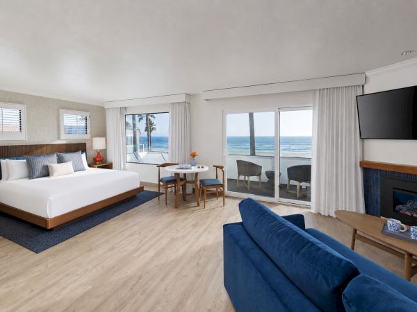 Modern beachfront hotel room with a king bed, blue sofa, fireplace, and large windows looking out to the ocean.