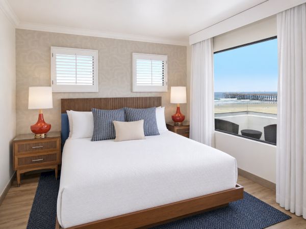 A modern seaside bedroom with a neatly made bed, lamps, and large windows showing the ocean and pier.